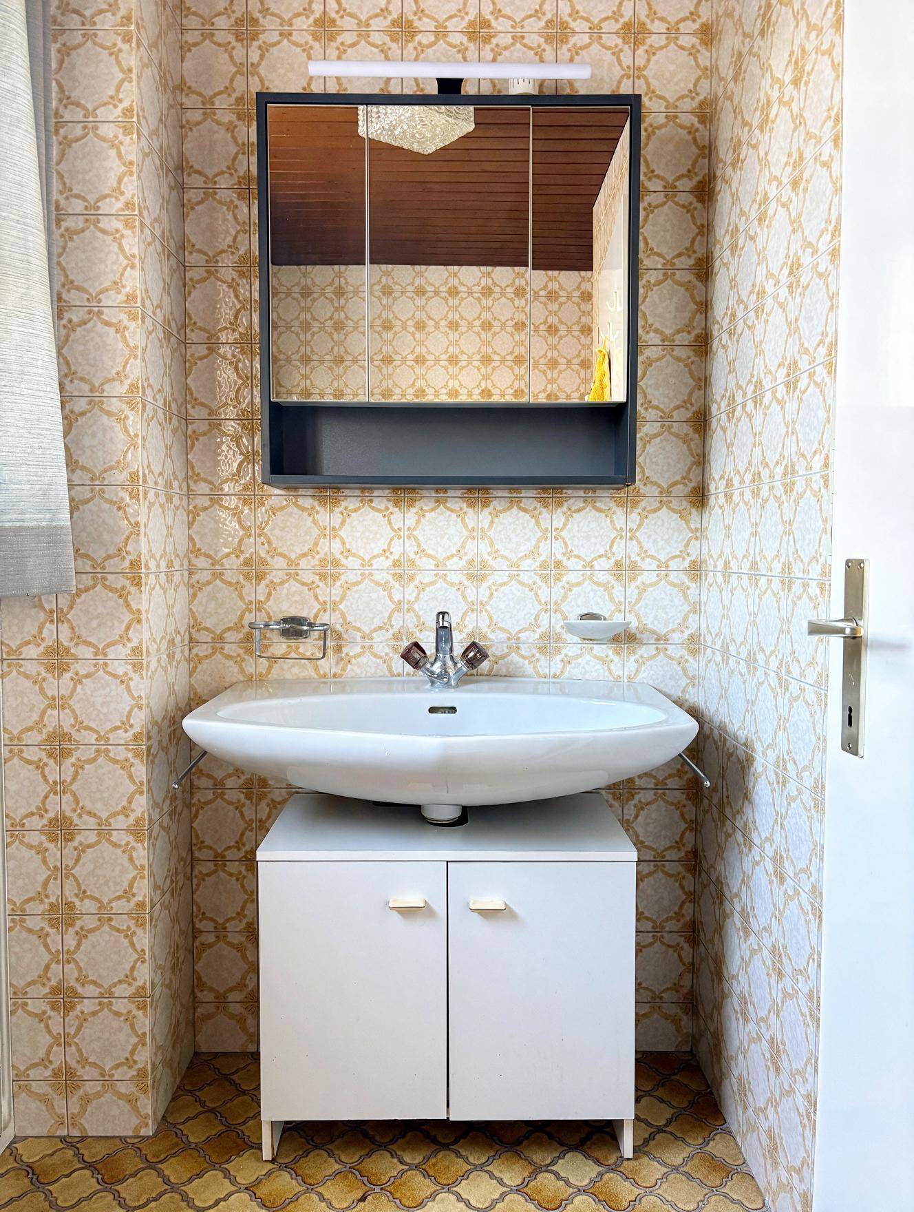 Badezimmer mit Waschbecken, Spiegelschrank und gemusterten Fliesen im charakteristischen 80er-Jahre-Stil.