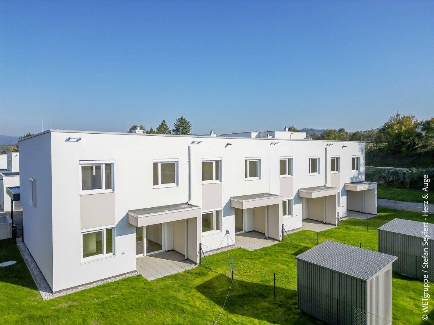 Helle Reihenhäuser mit privaten Gärten, Terrassen und Schuppen unter blauem Himmel.