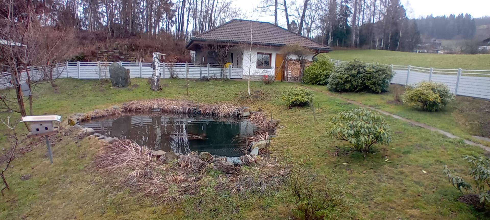 Großer Gartenbereich mit einem Teich und einem kleinen Nebengebäude, umgeben von einer Hecke.