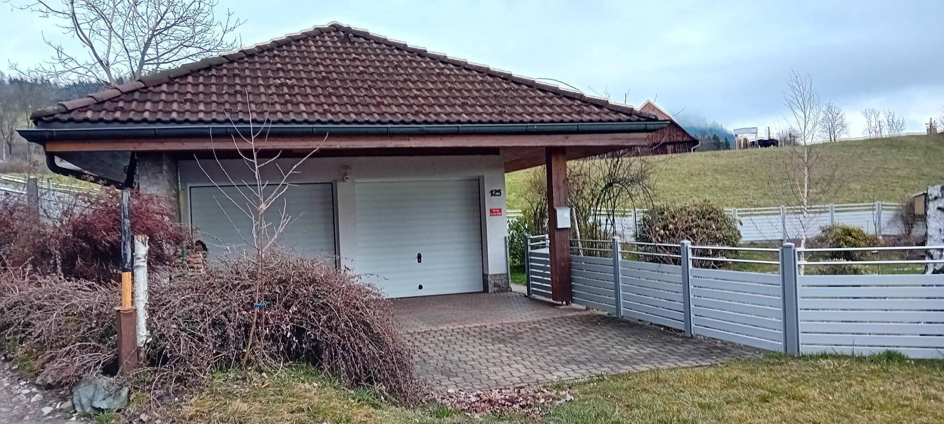 Einzelgarage mit Carport und gepflasterter Zufahrt, umgeben von grüner Landschaft.