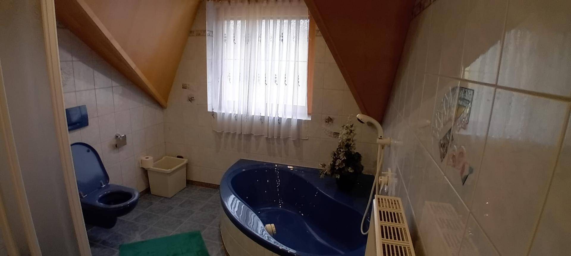 Badezimmer im Dachgeschoss mit blauer Eckbadewanne, WC und Fenster, unter einer Dachschräge.