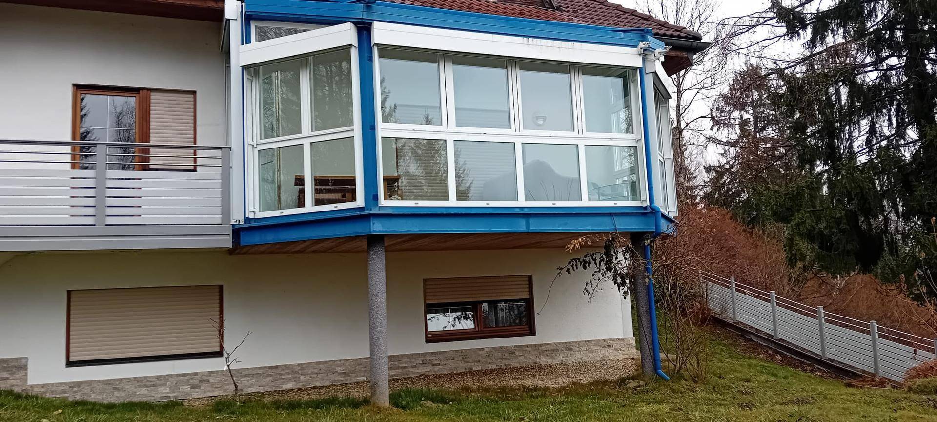 Detailansicht des blauen Wintergartens mit großen Fenstern und Rollläden am weißen Haus.