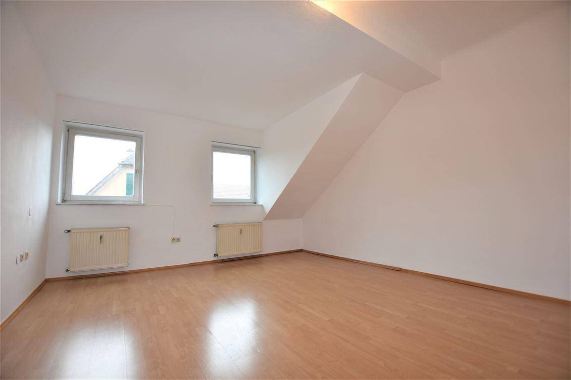 Leeres Zimmer mit Holzboden, weißen Wänden und zwei Fenstern unter einer Dachschräge.