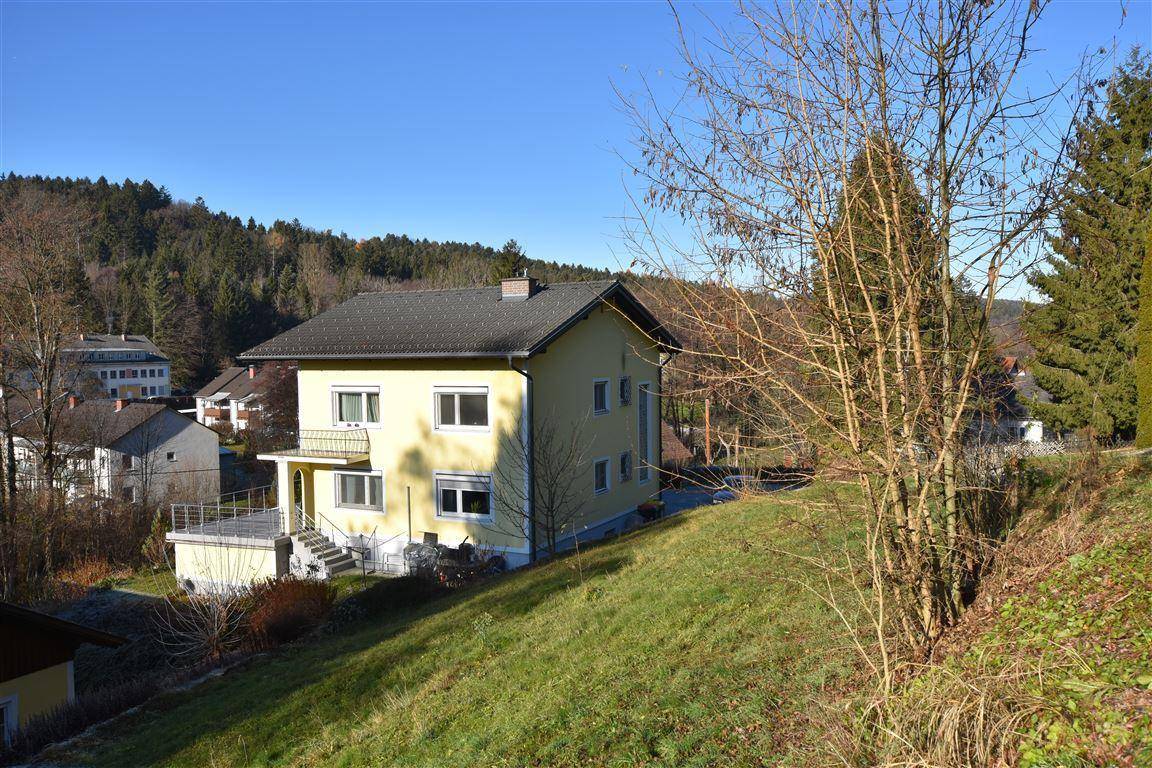 Gelbes Einfamilienhaus in Hanglage, umgeben von viel Grün und Bäumen unter blauem Himmel.