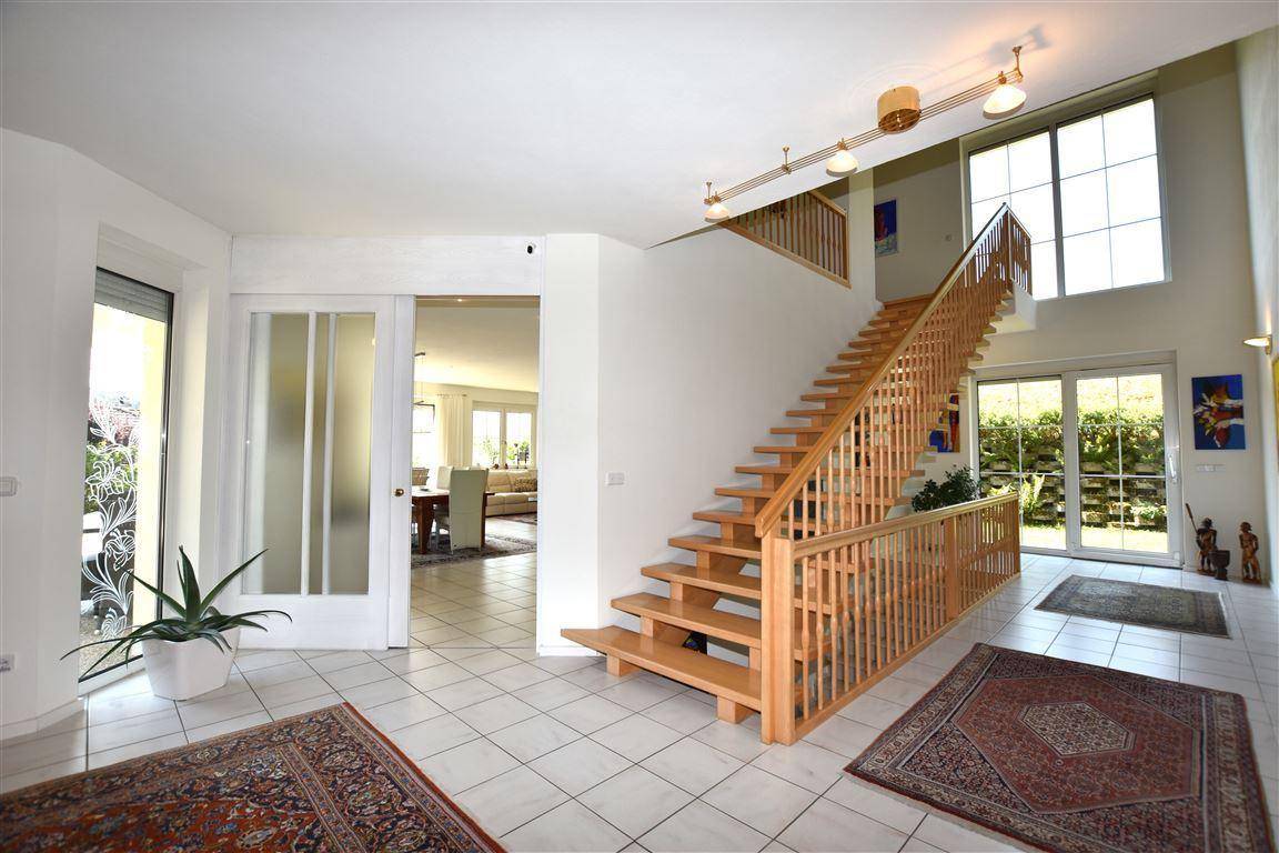 Geräumiger Eingangsbereich mit heller Holztreppe, Fliesenboden und viel Tageslicht durch große Fenster.