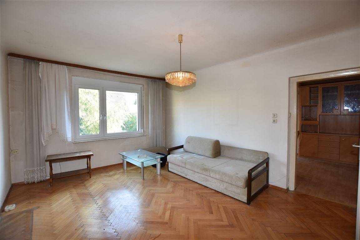 Wohnzimmer mit Fischgrätparkett, Sofa und Fenster mit Blick ins Grüne, angrenzend an einen weiteren Raum.