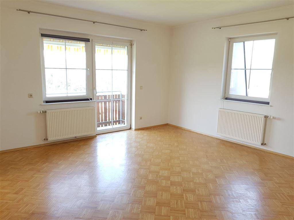 Geräumiges Wohnzimmer mit Parkettboden und zwei Fenstern, eines davon mit Balkonzugang.