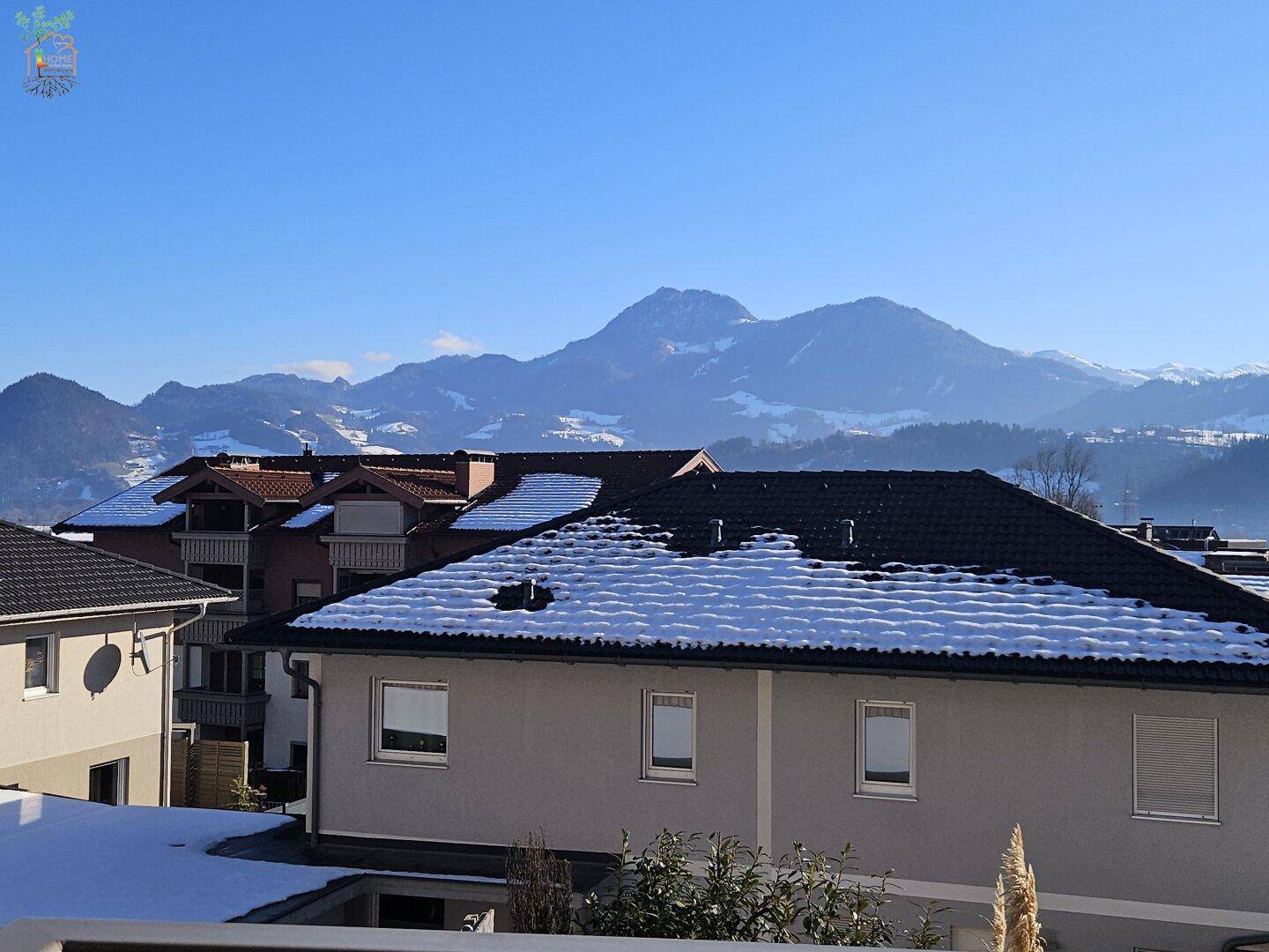 Winterliche Ansicht von Wohnhäusern mit schneebedeckten Dächern und majestätischen Bergen im Hintergrund.
