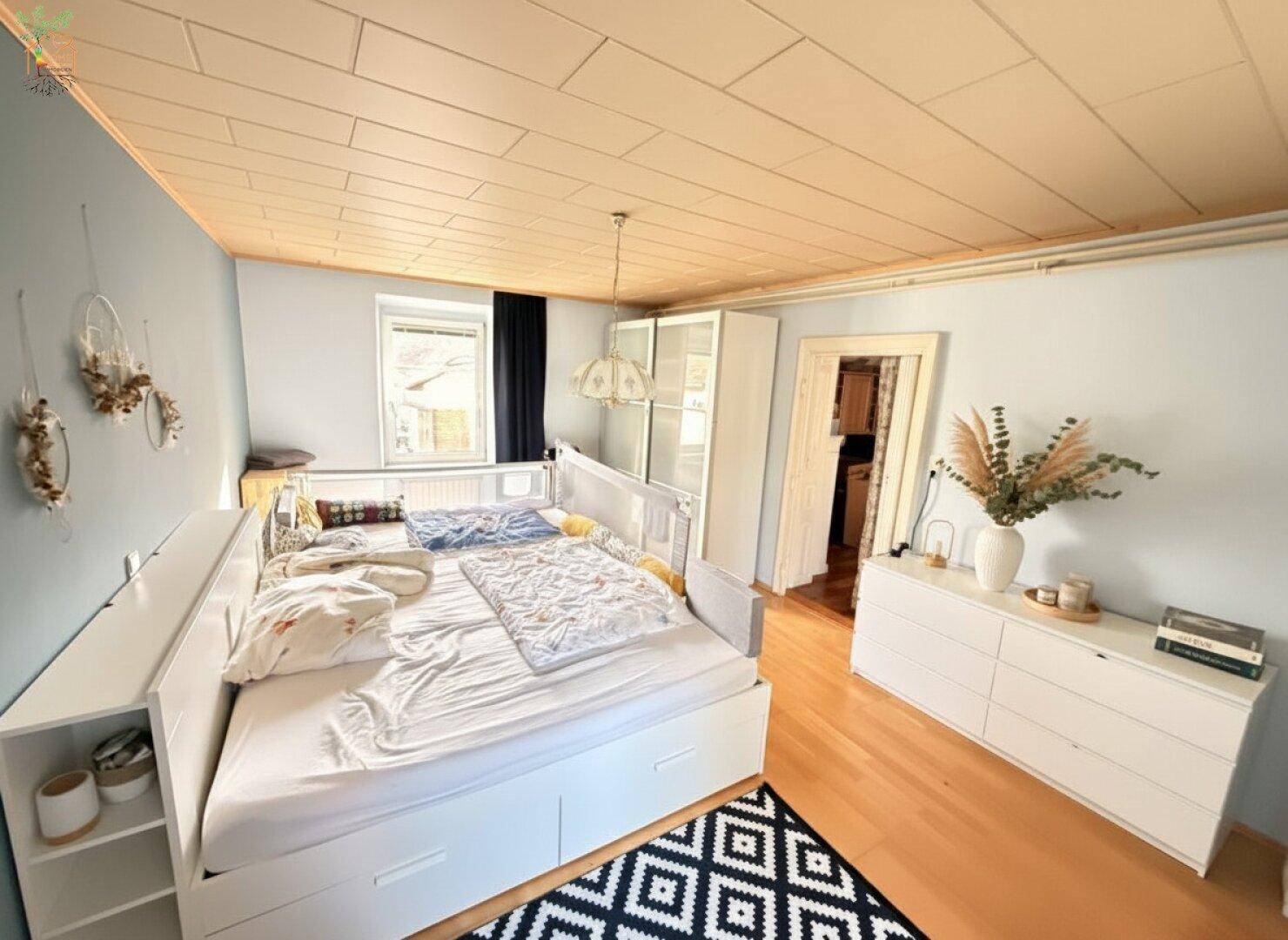 Helles Schlafzimmer mit großem Bett, Kommode und Holzdecke, freundlich gestaltet.