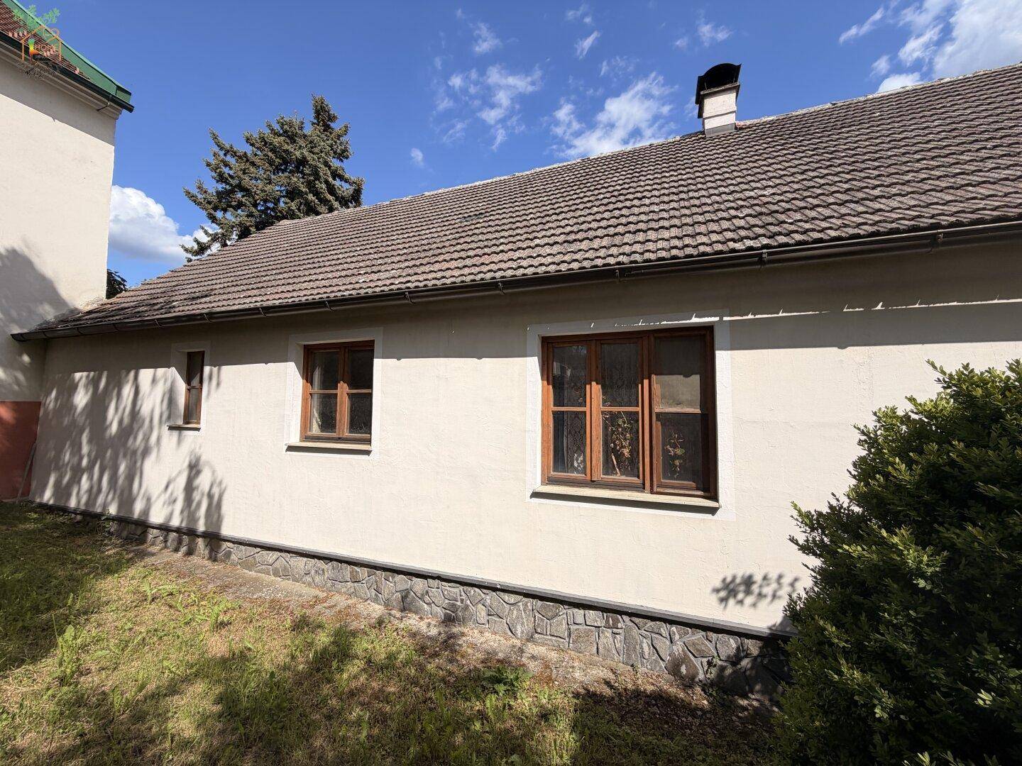 Seitenfassade des Hauses mit Steinsockel und Holzfenstern, umgeben von Grünflächen.