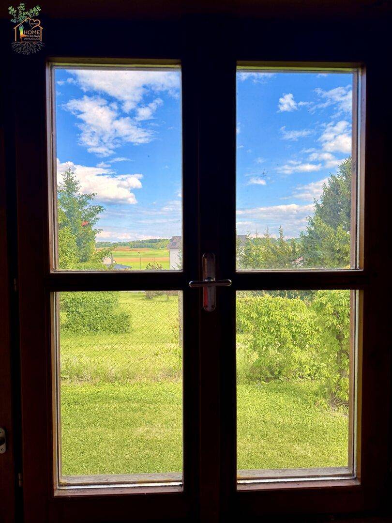 Blick aus dem Fenster auf den grünen Garten und die weite Landschaft unter blauem Himmel.