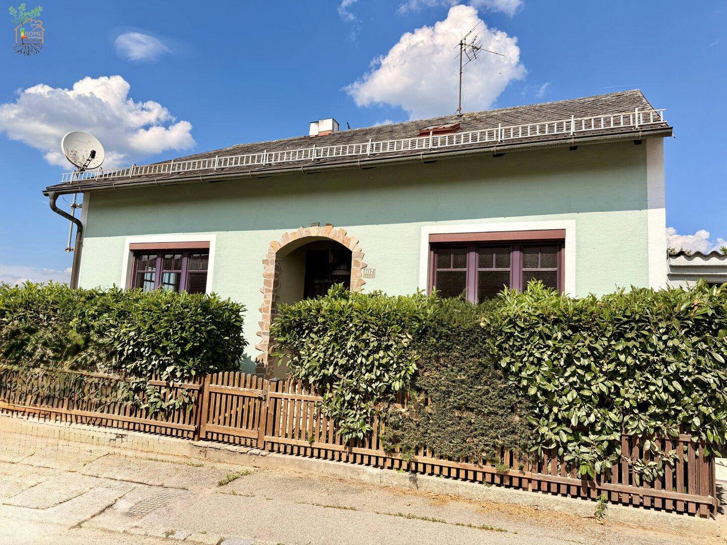 Vorderansicht des Hauses mit hellgrüner Fassade, Holzzaun und gepflegten Hecken.