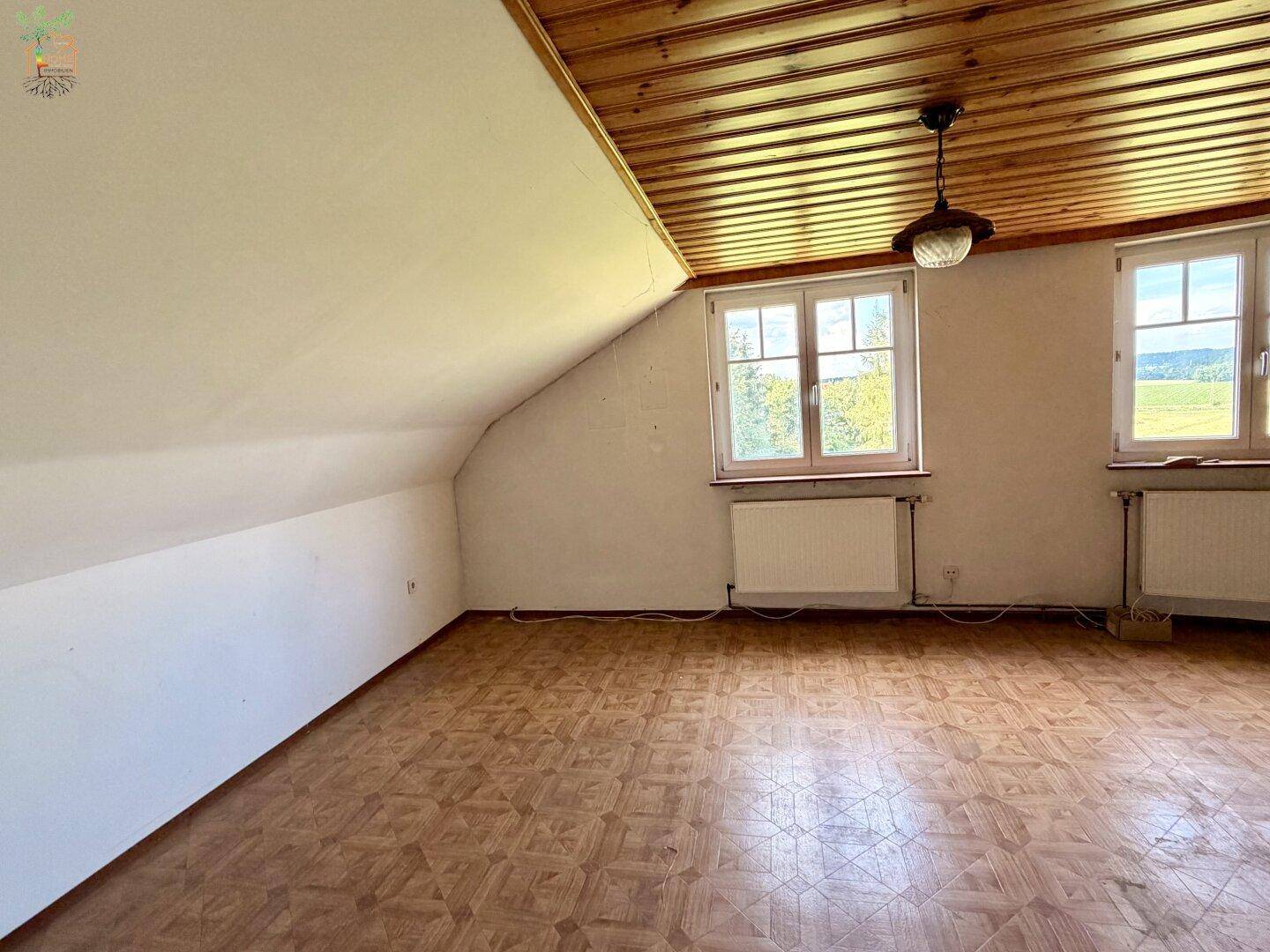 Helles Dachgeschosszimmer mit schrägen Wänden, Holzdecke und zwei Fenstern.