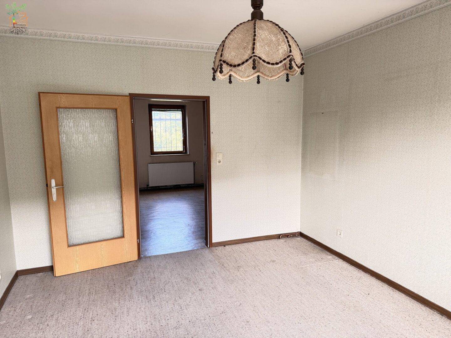Leeres Wohnzimmer mit Teppichboden, Zierleisten und Durchgang zu einem weiteren Raum.