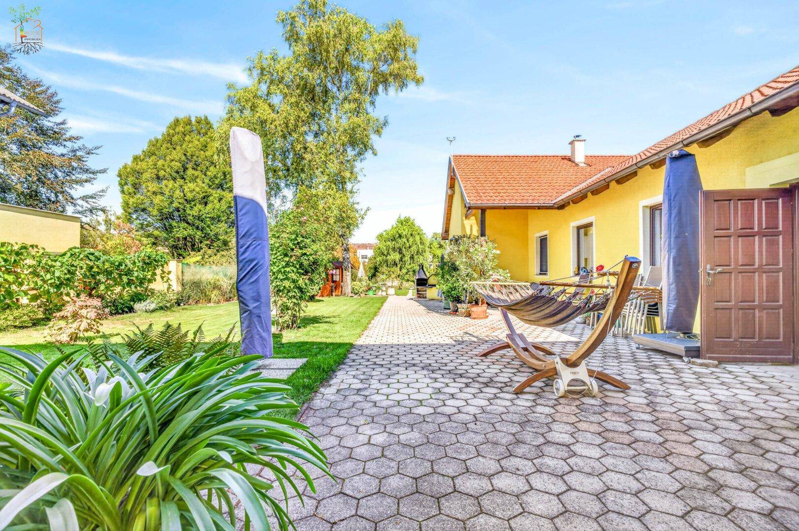 Weitläufiger Garten mit gepflasterter Terrasse, Hängematte und Grillbereich unter blauem Himmel.