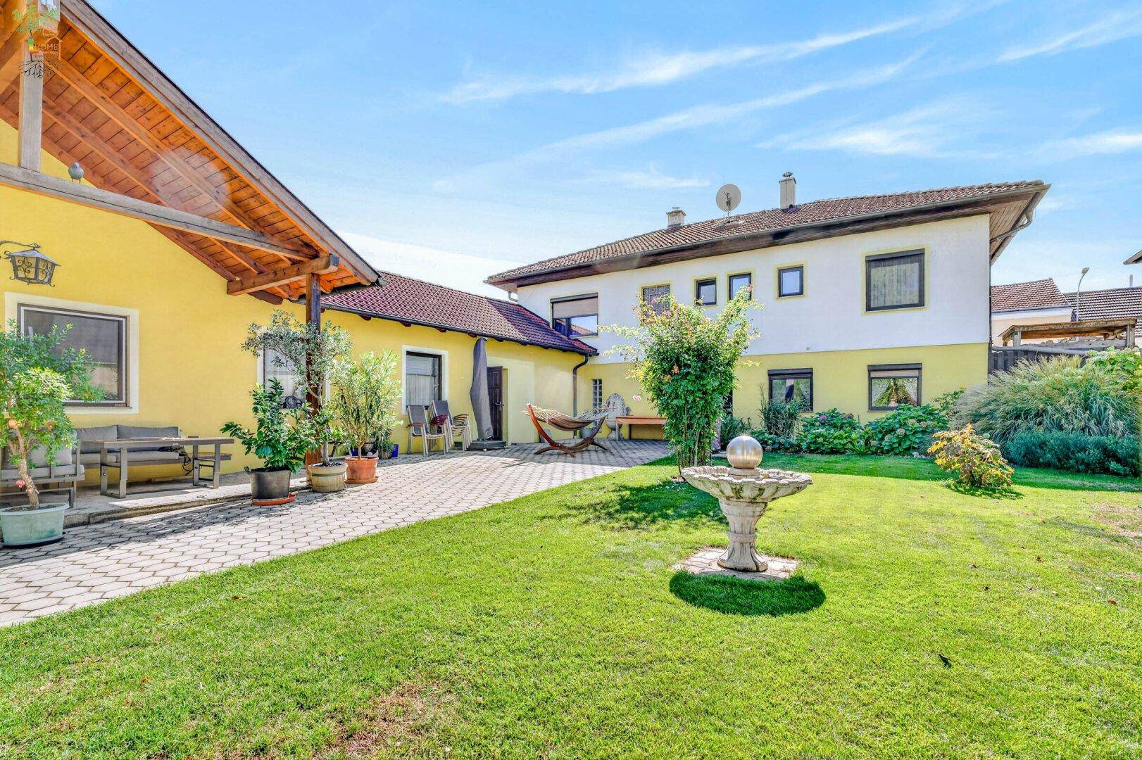 Großer Gartenbereich mit gepflasterter Terrasse, Rasenfläche und dekorativem Brunnen zwischen zwei Gebäuden.