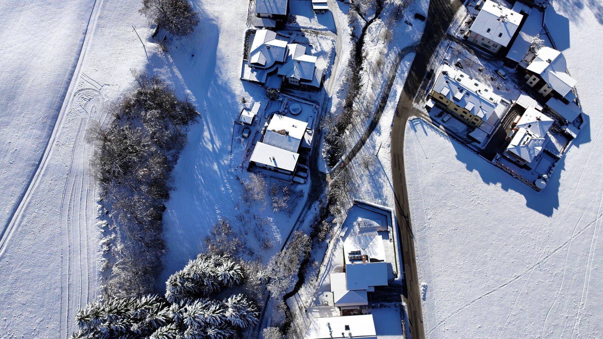 Luftaufnahme einer verschneiten Wohnsiedlung mit Häusern und Feldern in winterlicher Landschaft.
