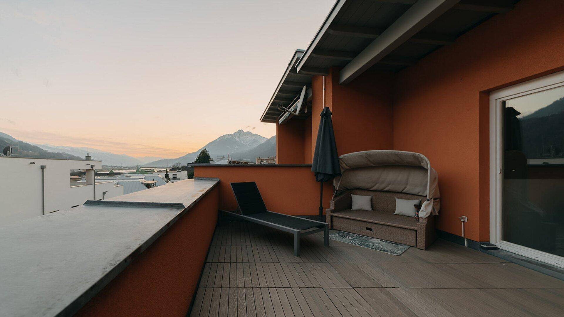 Geräumige Dachterrasse mit Holzbelag, Liegestuhl und Bergblick in der Abenddämmerung.