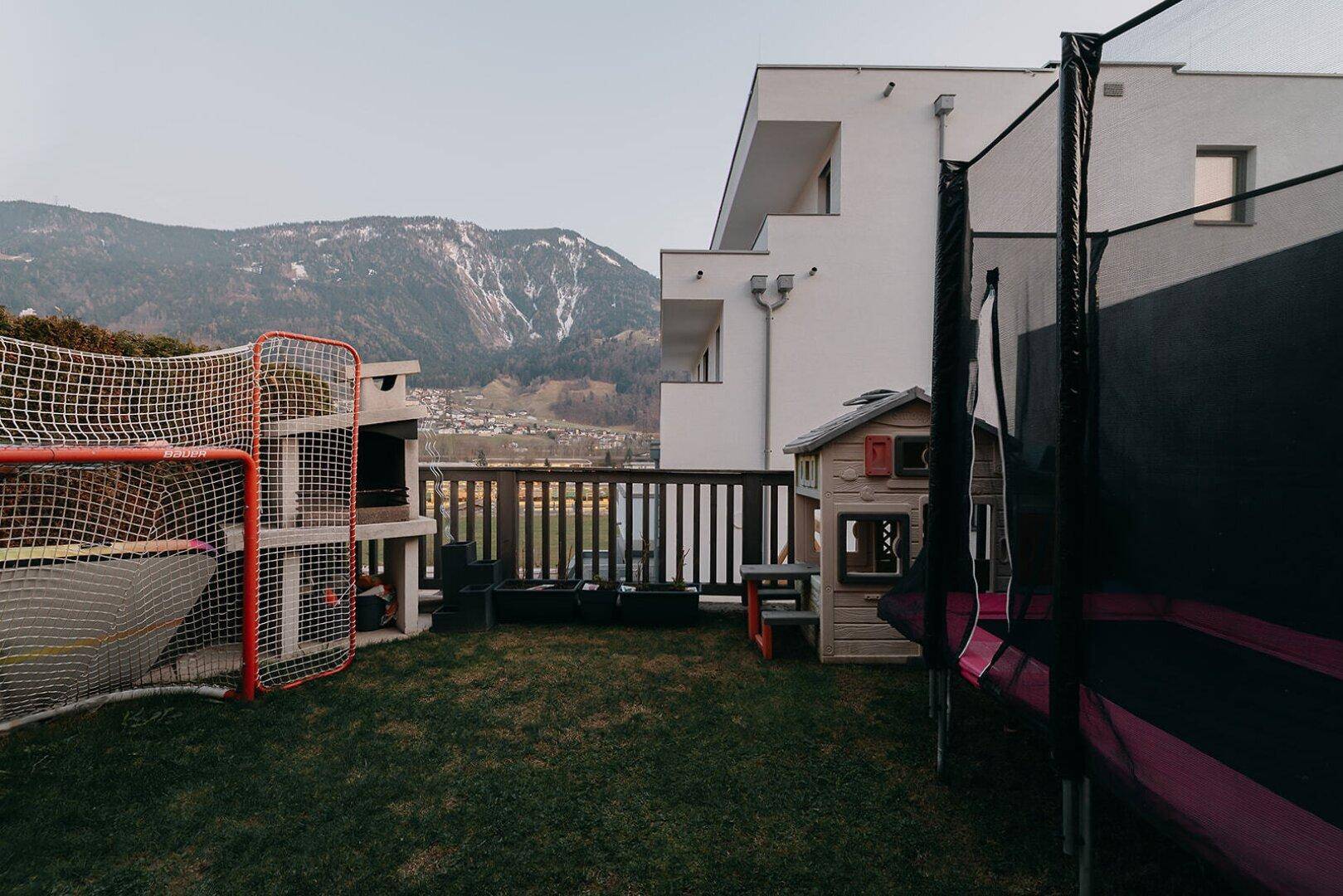 Garten mit Trampolin, Spielhaus und Grillplatz, umgeben von einer Bergkulisse.