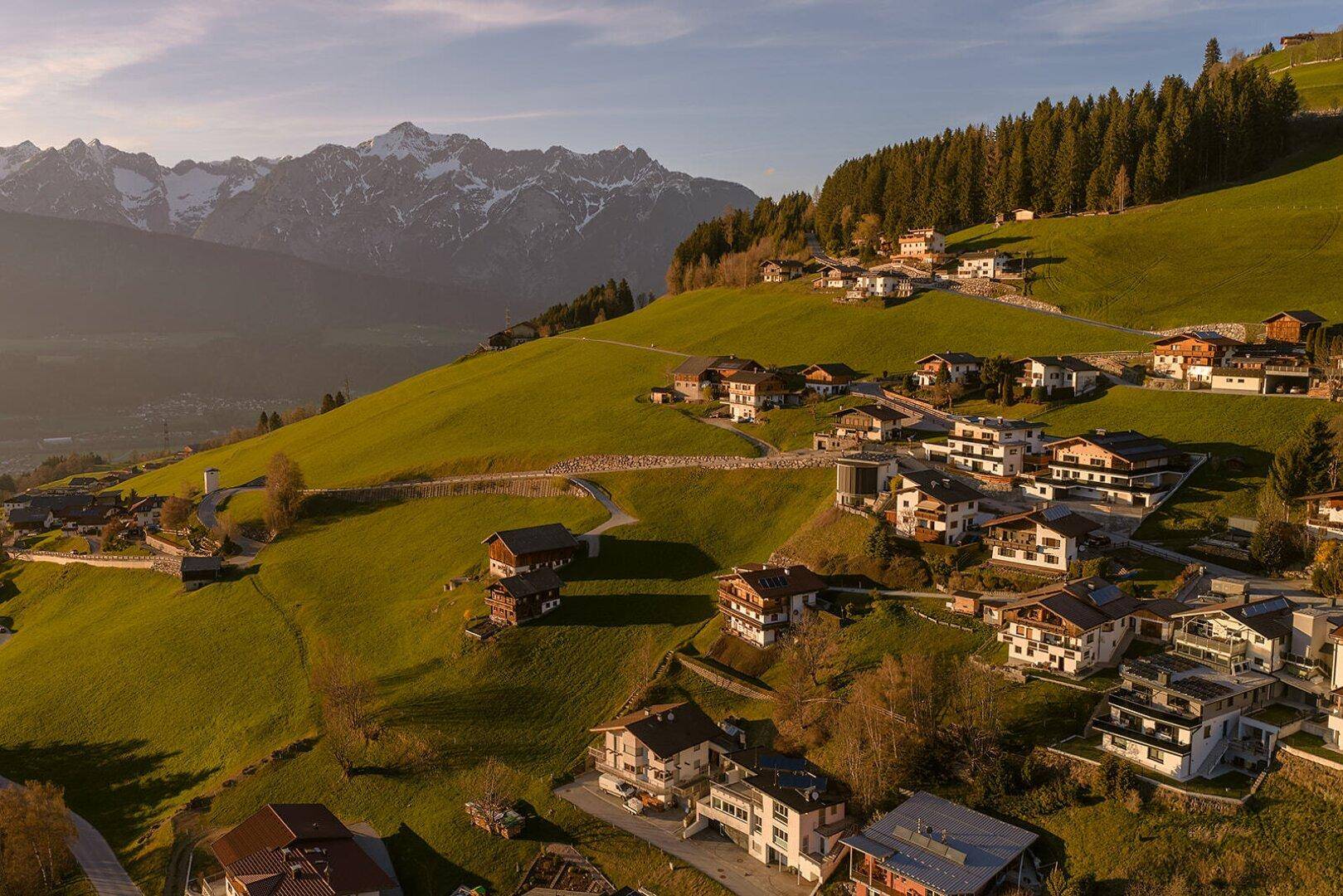 Weitläufige Ansicht einer Wohngegend auf einem grünen Hügel mit Waldrand und beeindruckender Bergkulisse.