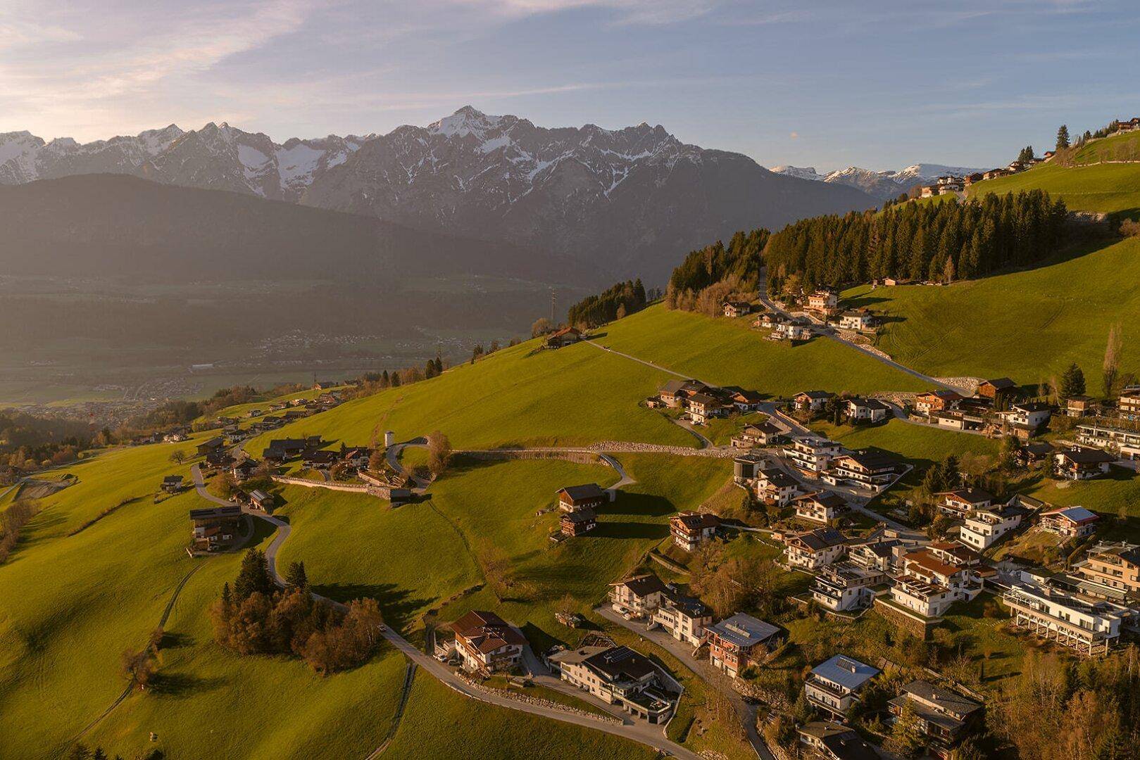 Panoramablick auf ein Dorf in den Alpen, eingebettet in grüne Hügel mit schneebedeckten Bergen im Hintergrund.