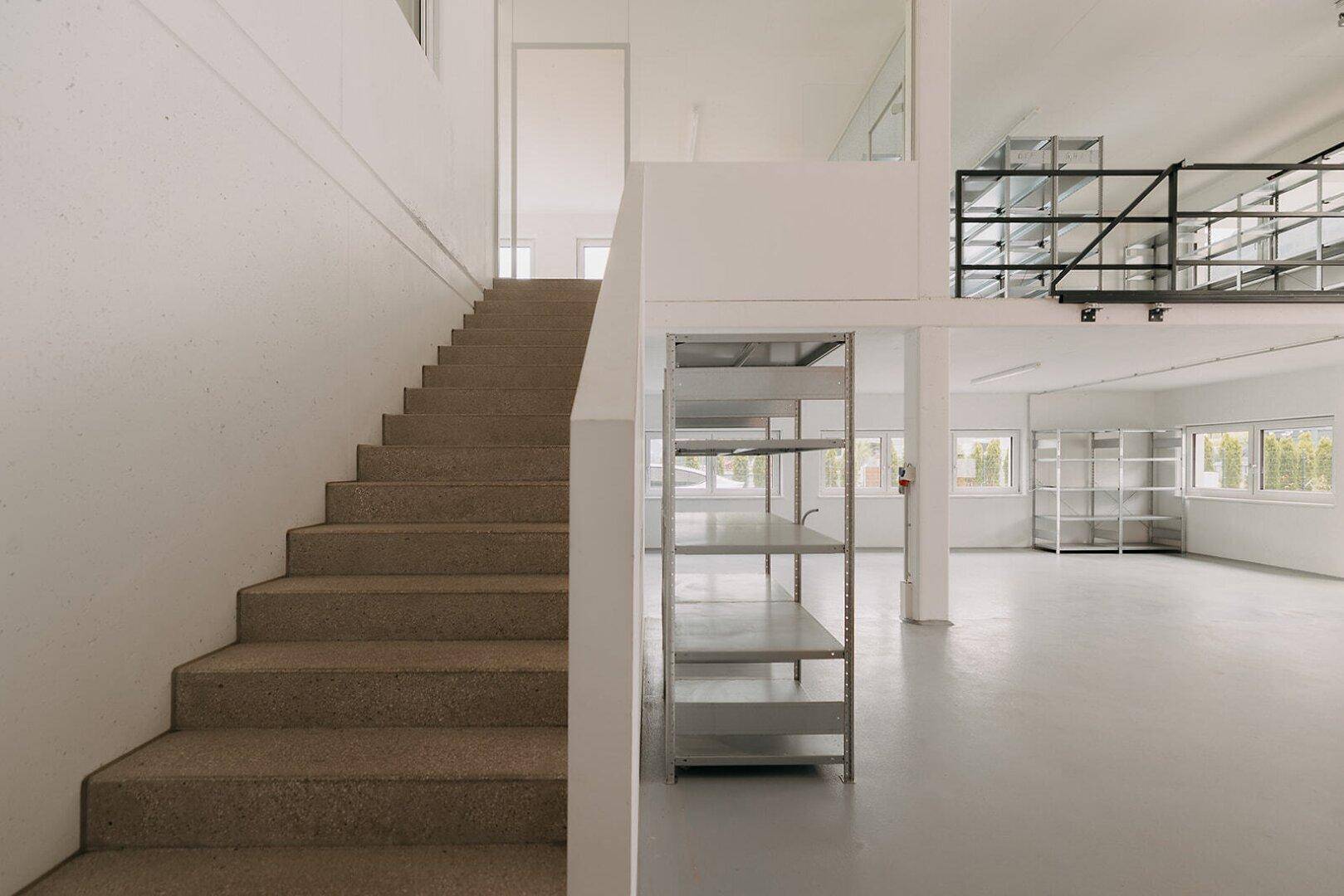 Treppenaufgang zu einem Zwischengeschoss in einer hellen Lagerhalle mit Betonboden.