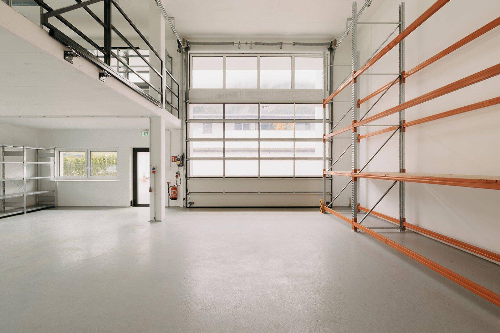 Geräumige Lagerhalle mit Betonboden, Rolltor und einem Zwischengeschoss mit Metallgeländer.