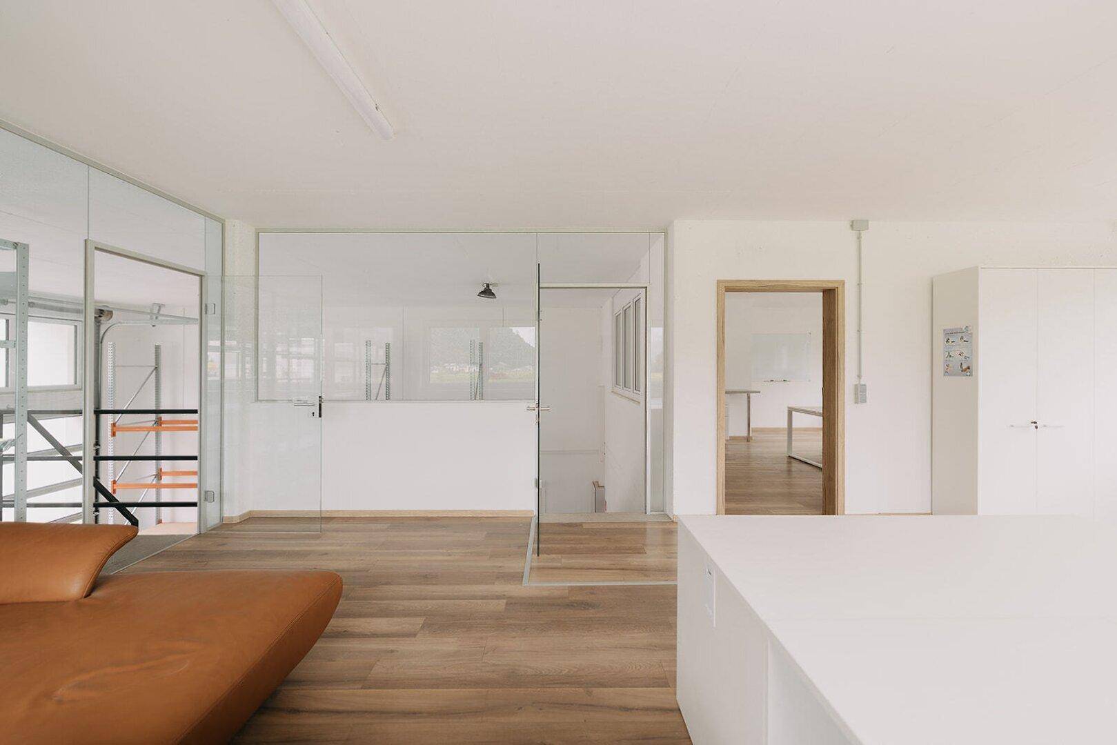 Moderner Bürobereich mit Holzboden, Ledersofa und Glaswänden, die den Raum unterteilen.