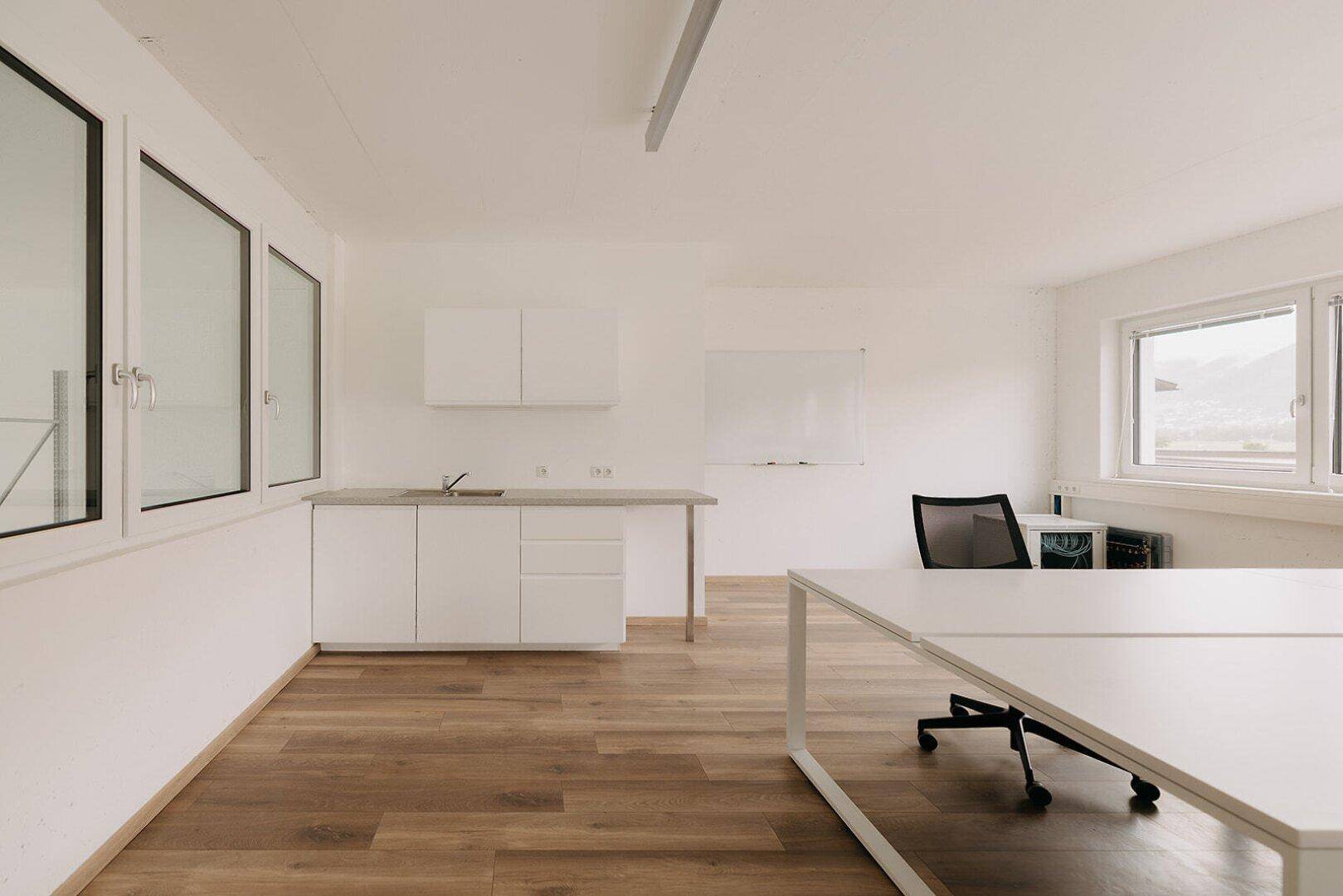 Modernes Büro mit Holzboden, integrierter Küchenzeile und großen Fenstern für viel Tageslicht.