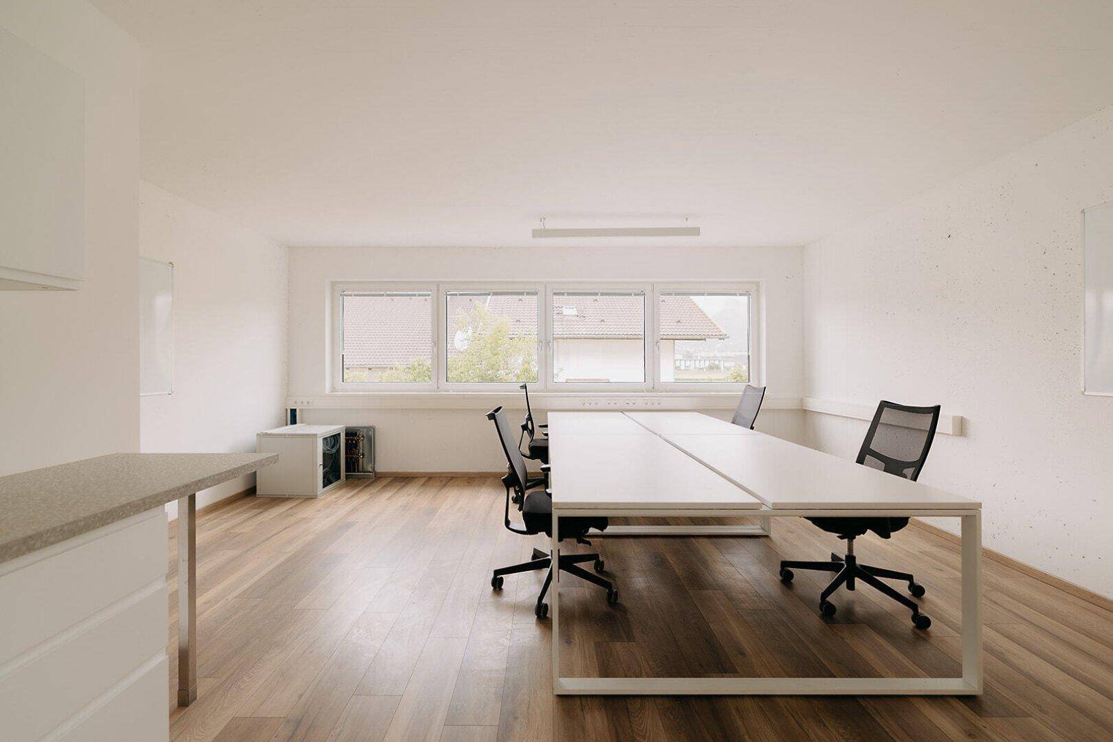 Heller Bürobereich mit Holzboden, modernen Schreibtischen und Fenstern mit Blick ins Grüne.