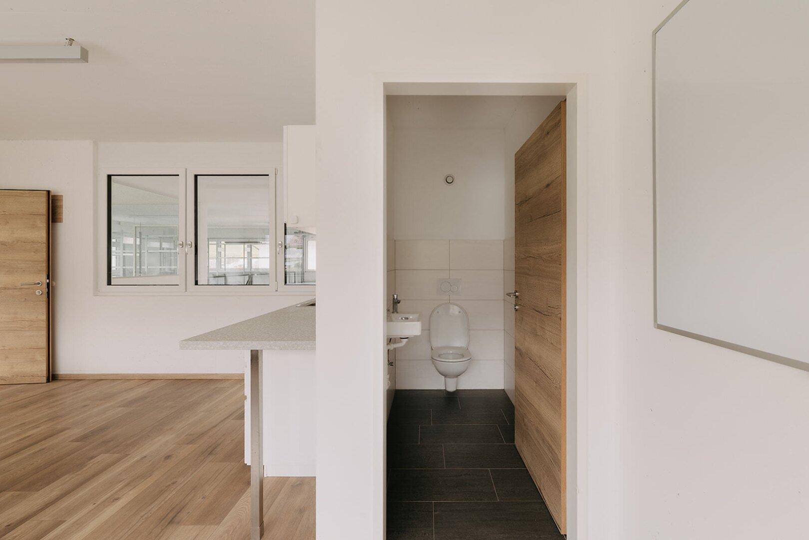 Kleiner, sauberer Toilettenraum mit WC, Waschbecken und dunklen Fliesen, zugänglich vom Bürobereich.