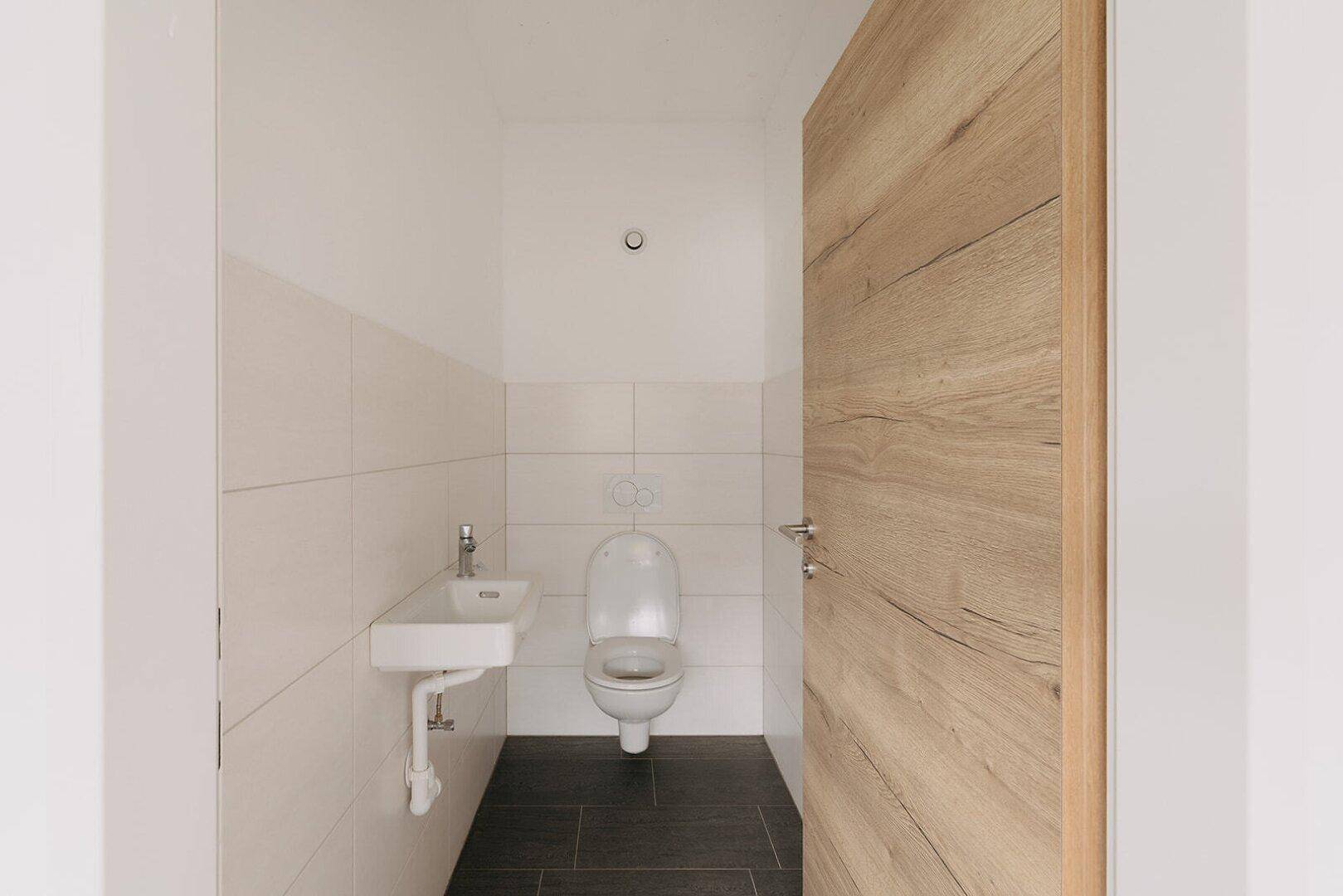 Detailansicht eines gepflegten Gäste-WCs mit Wand-WC, kleinem Waschbecken und hellen Wandfliesen.
