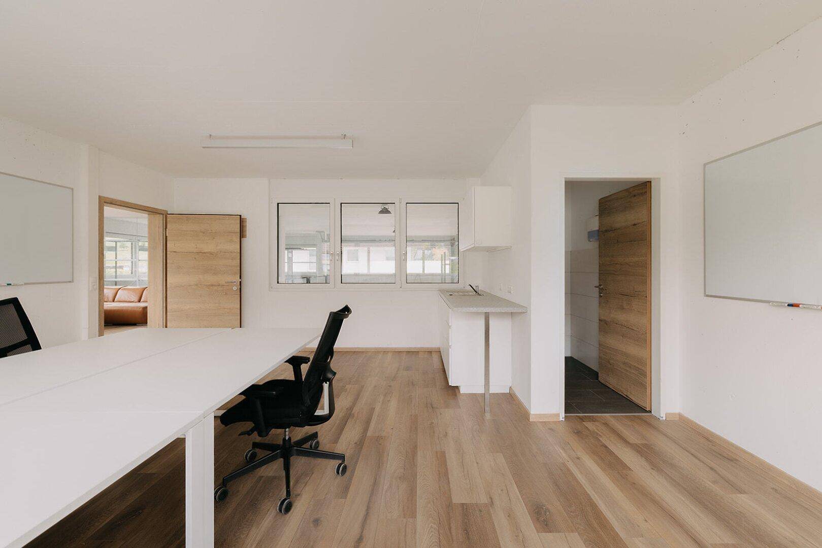 Heller und moderner Büroraum mit Holzboden, großen Fenstern und funktionalen Arbeitsplätzen.