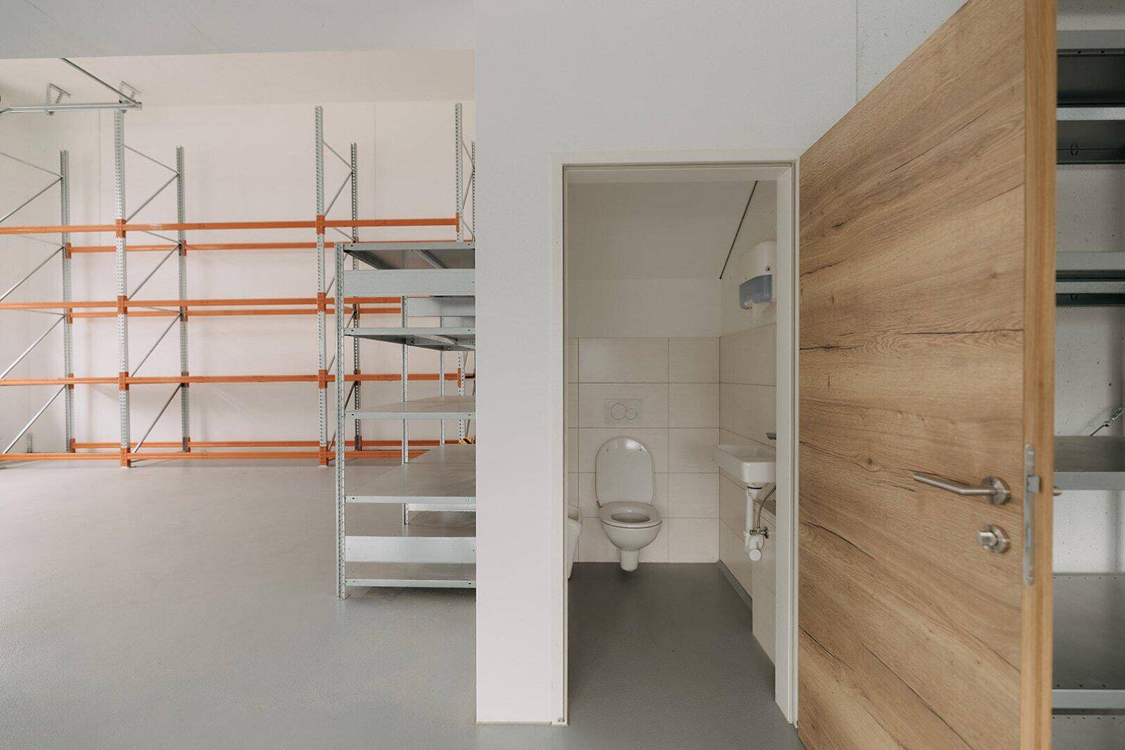Blick vom Lagerbereich in einen kleinen Toilettenraum mit WC und Waschbecken, umgeben von Regalen.