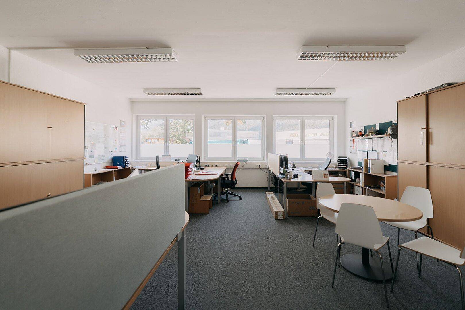 Möbliertes Büro mit mehreren Arbeitsplätzen, Teppichboden und großen Fenstern für natürliches Licht.