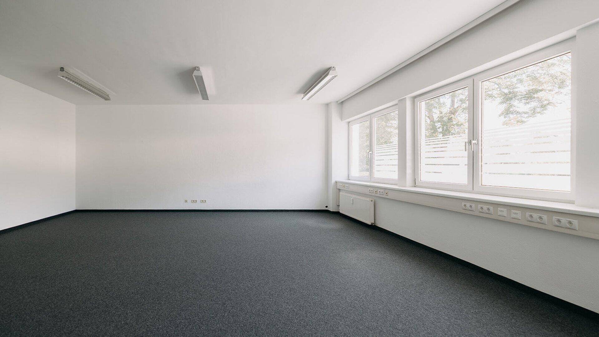 Helles, leeres Büro mit Teppichboden und einer Reihe von Fenstern, die viel Tageslicht hereinlassen.