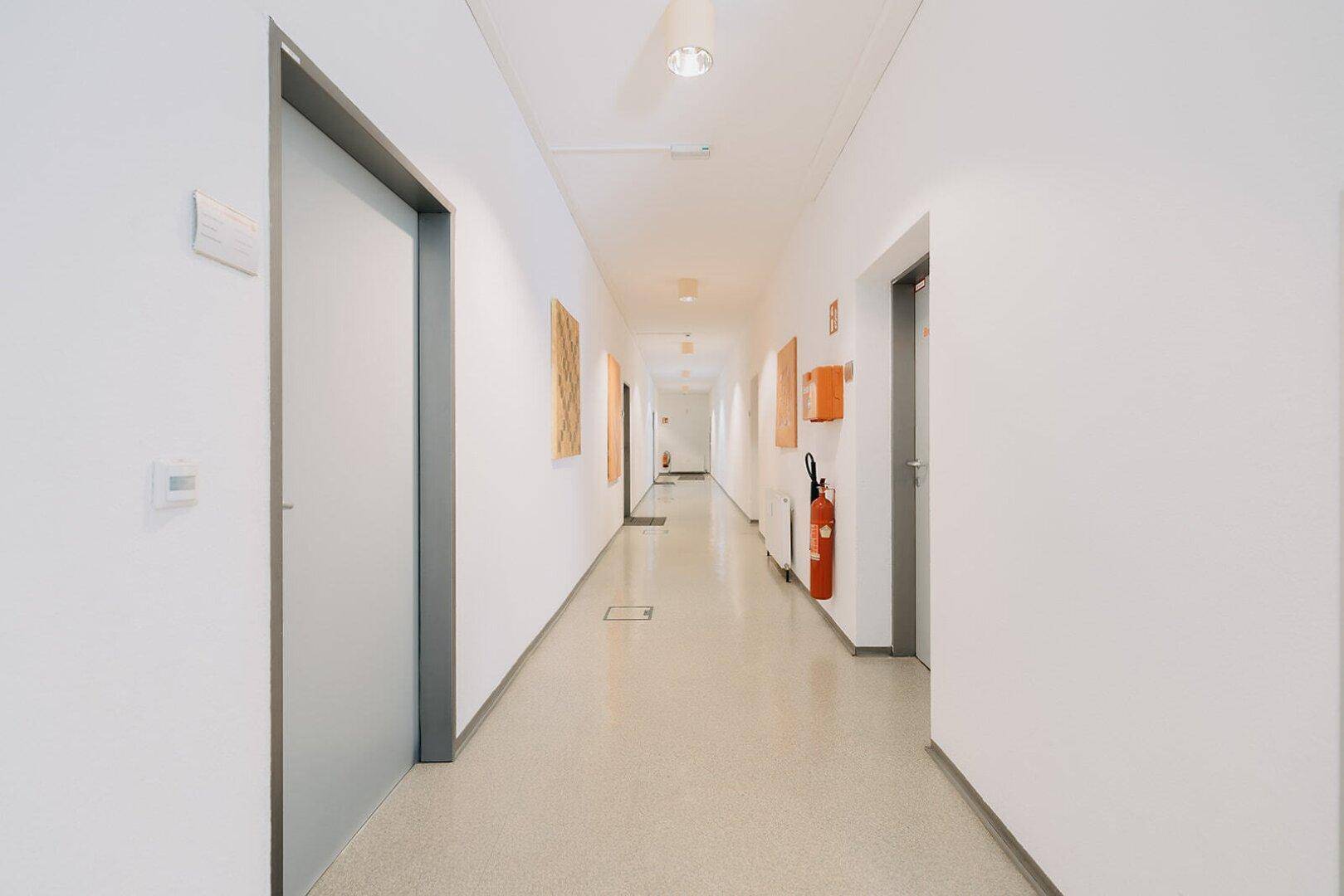 Langer, heller Flur mit Türen zu verschiedenen Räumen und einem Feuerlöscher an der Wand.