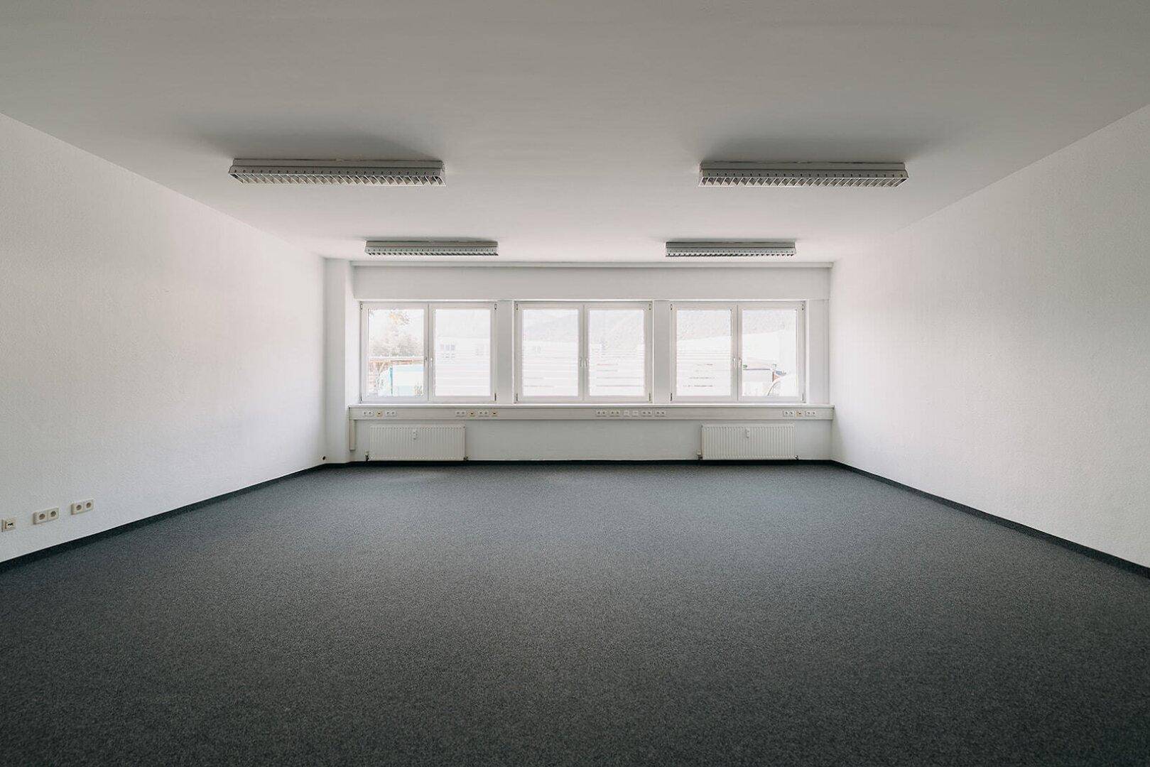 Heller Büroraum mit Teppichboden und einer Fensterfront, die für gute Belichtung sorgt.