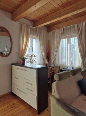 Gemütlicher Wohnbereich mit Holzbalkendecke, hellen Vorhängen und einem Sideboard mit Spiegel.