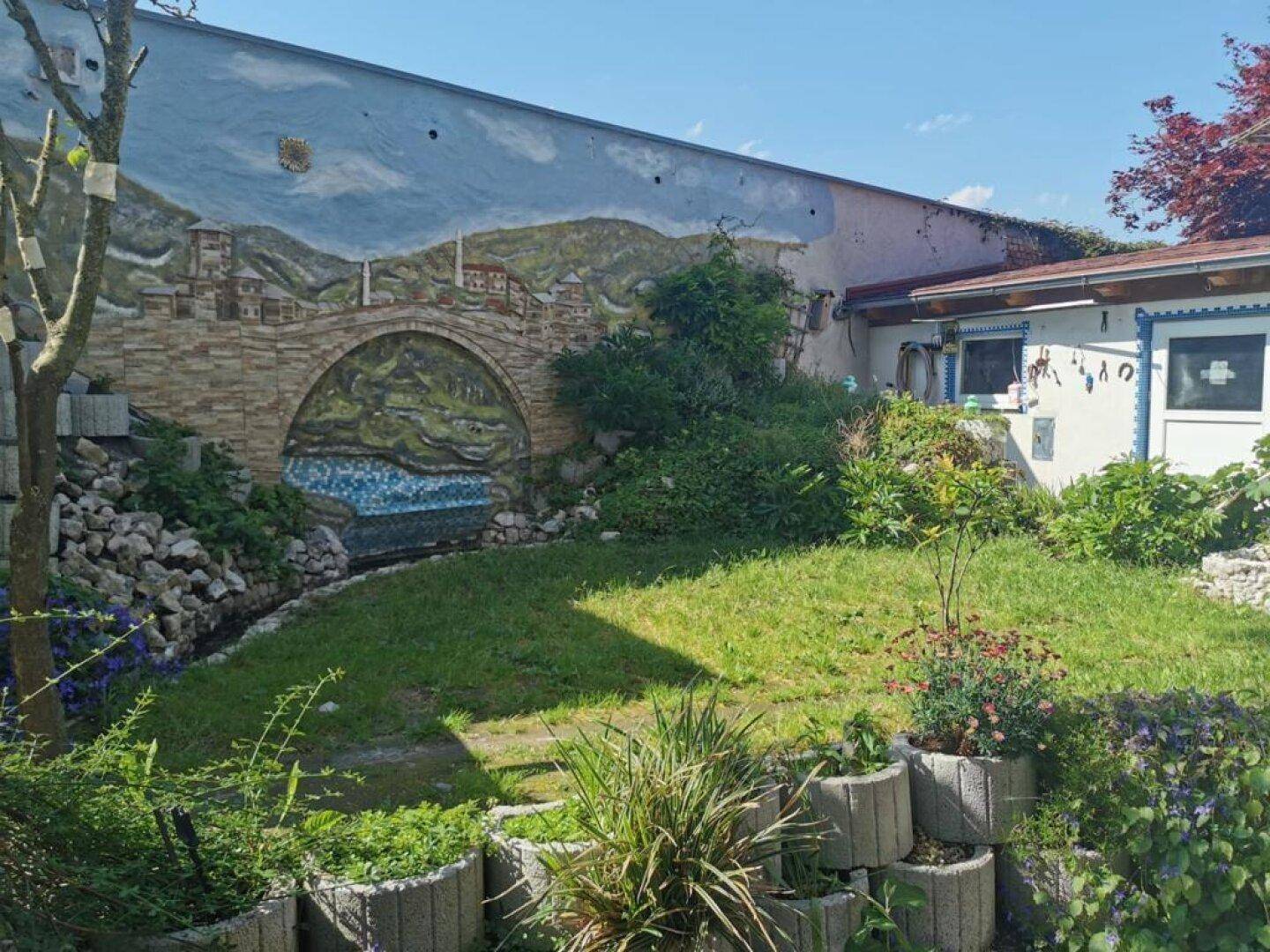 Grüner Gartenbereich mit einer bemalten Wand, die eine Brückenlandschaft darstellt, und verschiedenen Pflanzen.