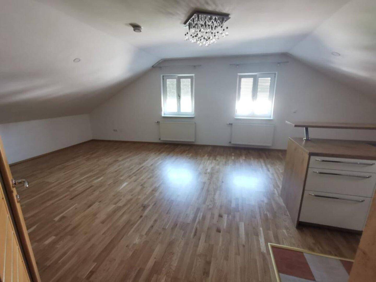 Geräumiger Wohnbereich mit Holzboden, Dachschrägen und zwei Fenstern, die viel Licht spenden.