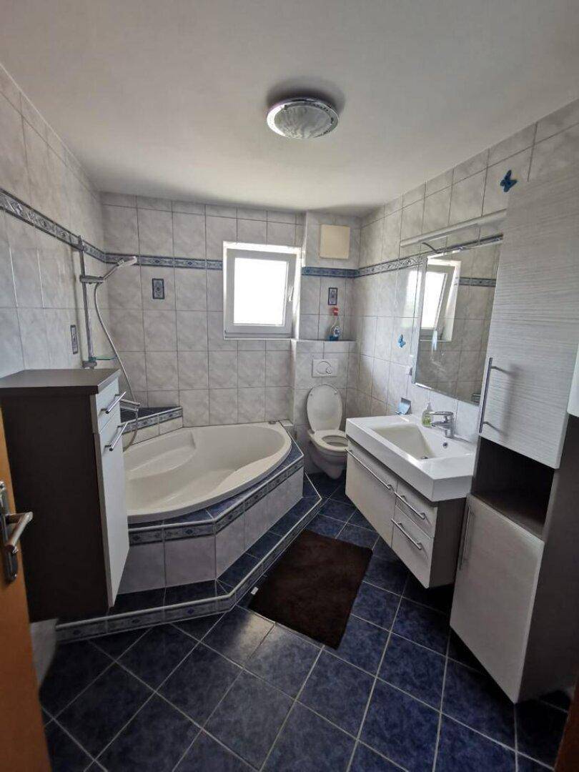 Badezimmer mit Eckbadewanne, blauen Bodenfliesen und einem Fenster für Belüftung und Licht.