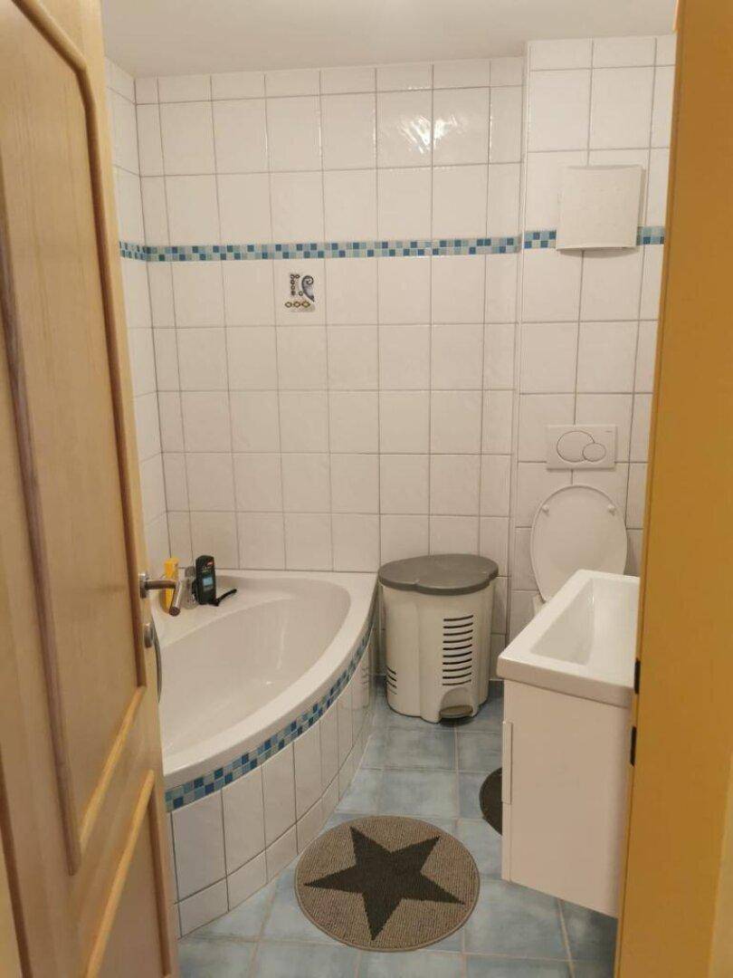 Badezimmer mit Eckbadewanne, weißen Fliesen und einem kleinen Waschbeckenunterschrank.
