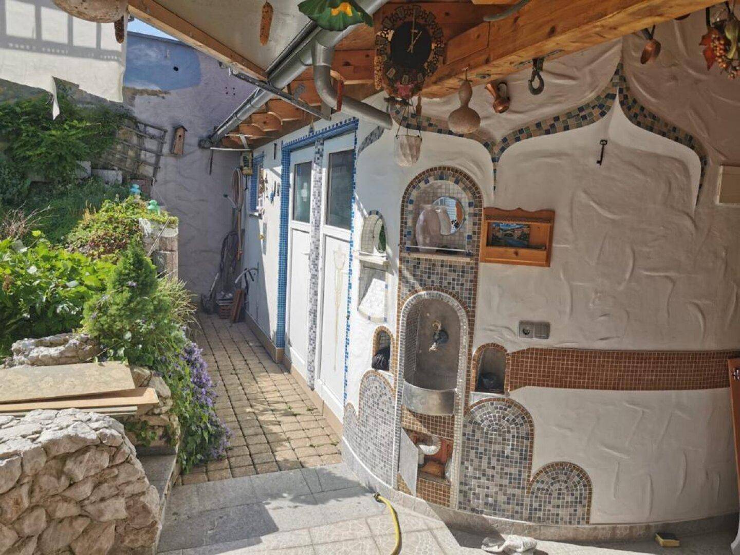 Außenbereich mit gepflastertem Weg, dekorativen Mosaikdetails an der Wand und üppiger Bepflanzung.