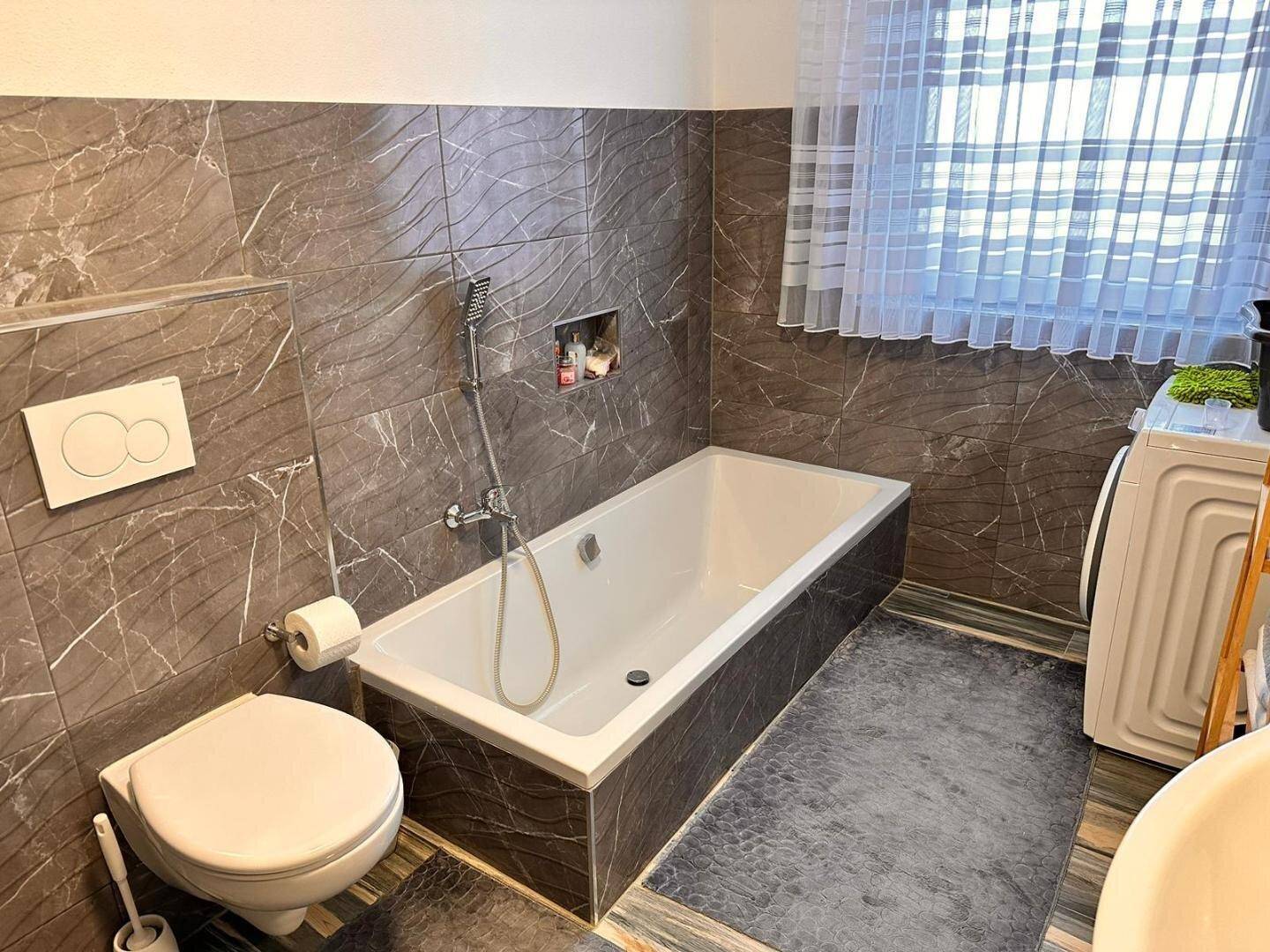 Badezimmer mit Badewanne, Wand-WC und Waschmaschine, ausgestattet mit dunklen Fliesen.