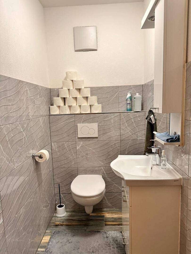 Gäste-WC mit Wand-WC, Waschbecken und grauen Fliesen, inklusive praktischem Spiegelschrank.