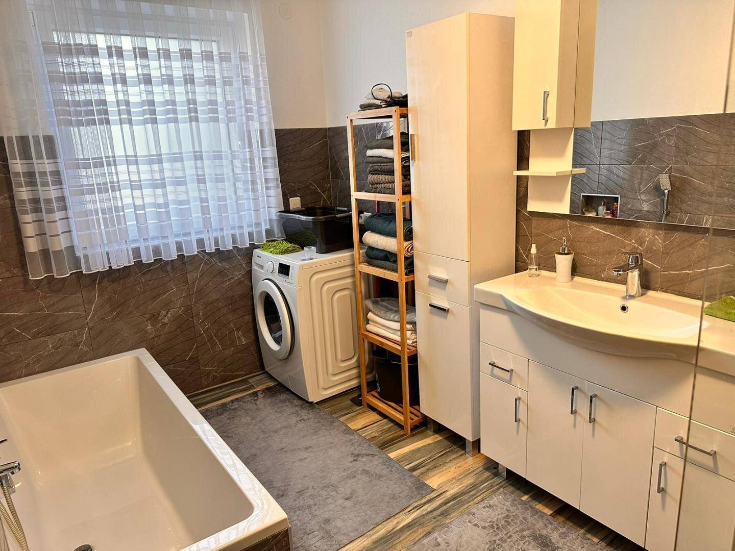 Helles Badezimmer mit Waschbecken, Badewanne und Waschmaschine, gut beleuchtet durch ein Fenster.