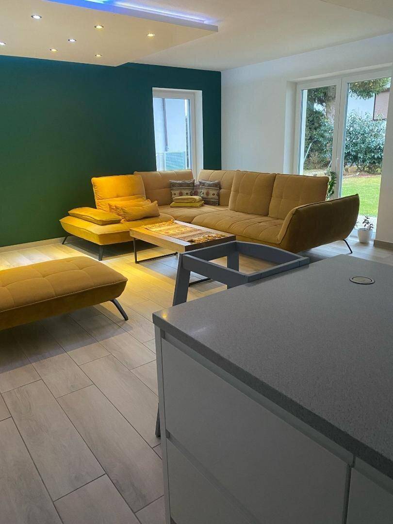 Heller Wohnbereich mit gelbem Ecksofa, Holzboden und einer Akzentwand in Dunkelgrün.