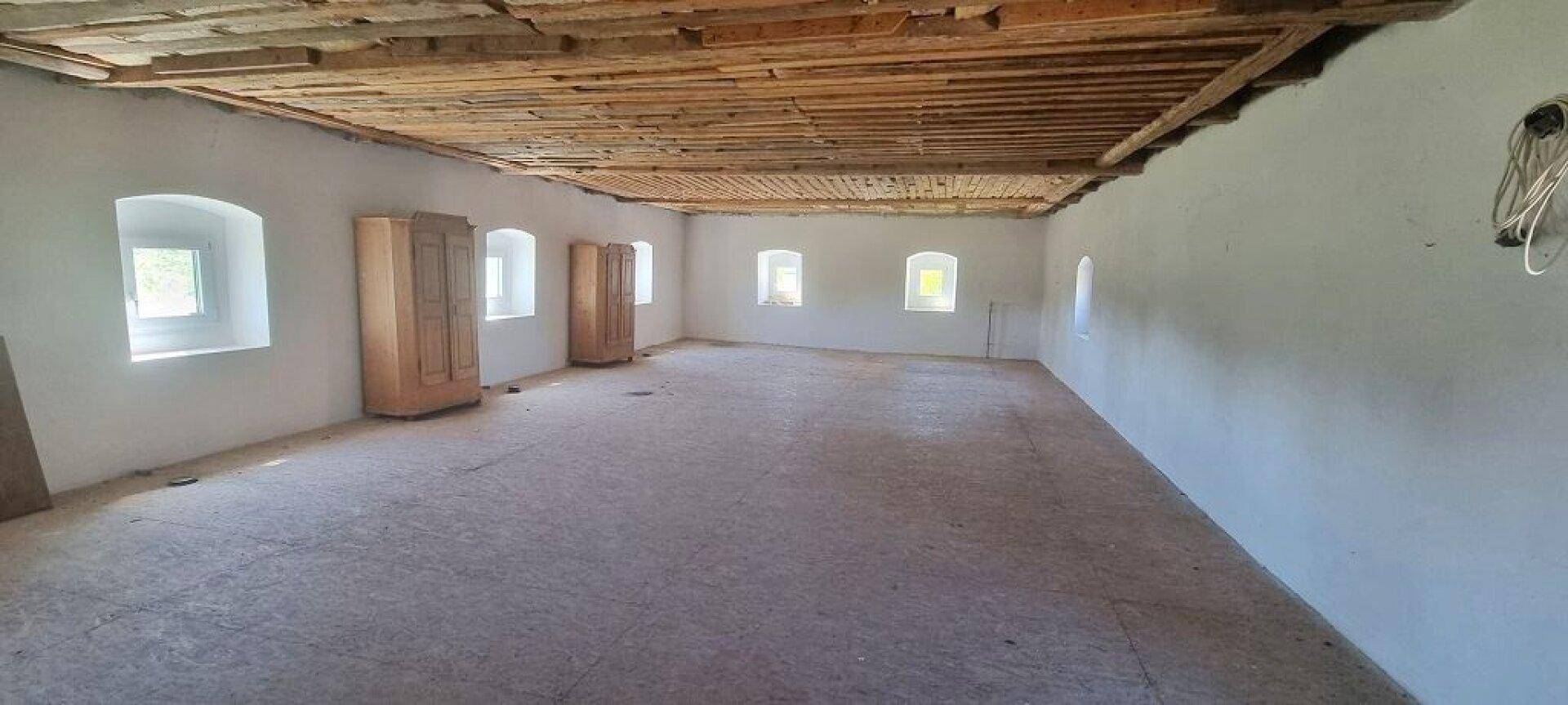 Leerer Raum mit Betonboden, weißen Wänden und sichtbaren Holzdeckenbalken.