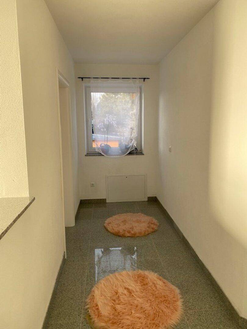 Schmaler Flur mit einem Fenster, das Tageslicht hereinlässt, und dekorativen Teppichen auf dem Fliesenboden.